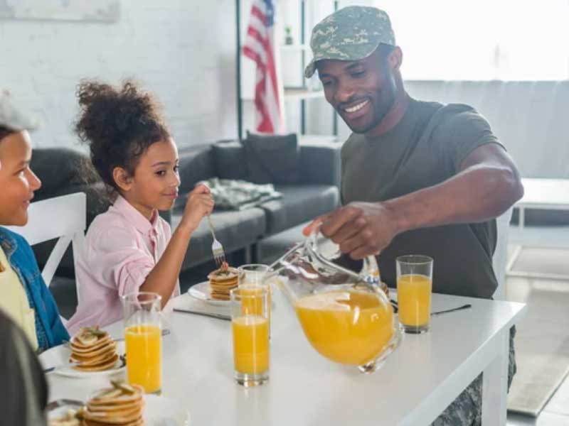 A man in a military uniform serves pancakes to a young girl at a family breakfast table.