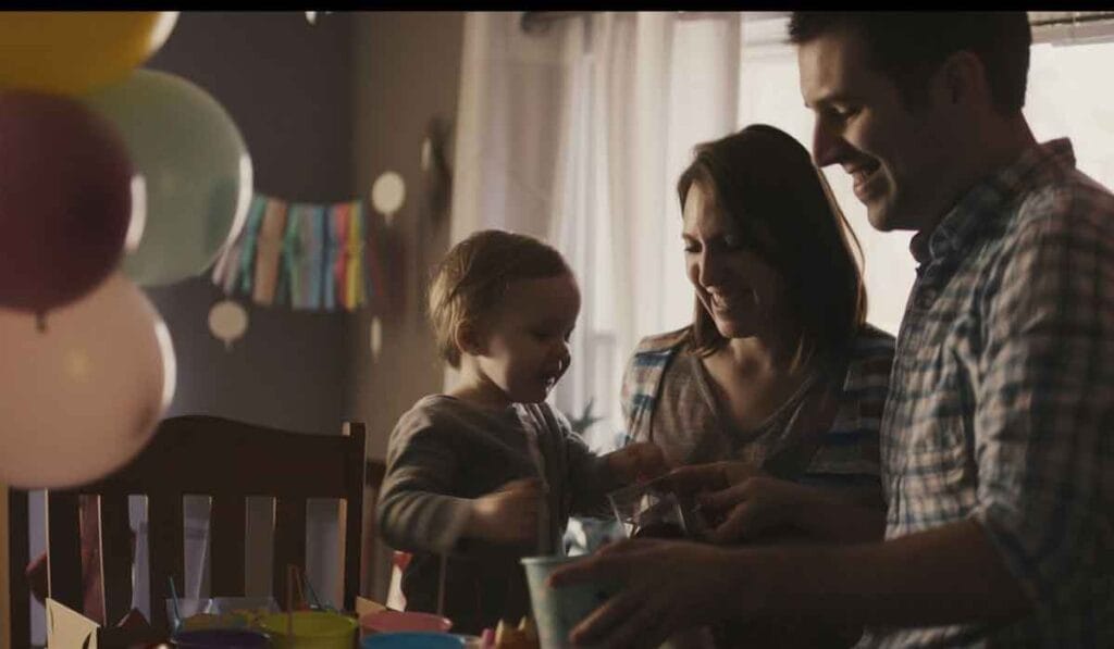 A family of three, with a child and two adults, are gathered at a table with colorful balloons and decorations. They appear to be celebrating a special occasion.