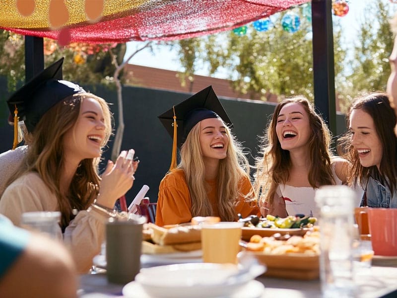 Group of young adults celebrating at an outdoor event, some wearing graduation caps. They are sitting around a table in Tempe, AZ filled with food, laughing and enjoying the sunny weather.